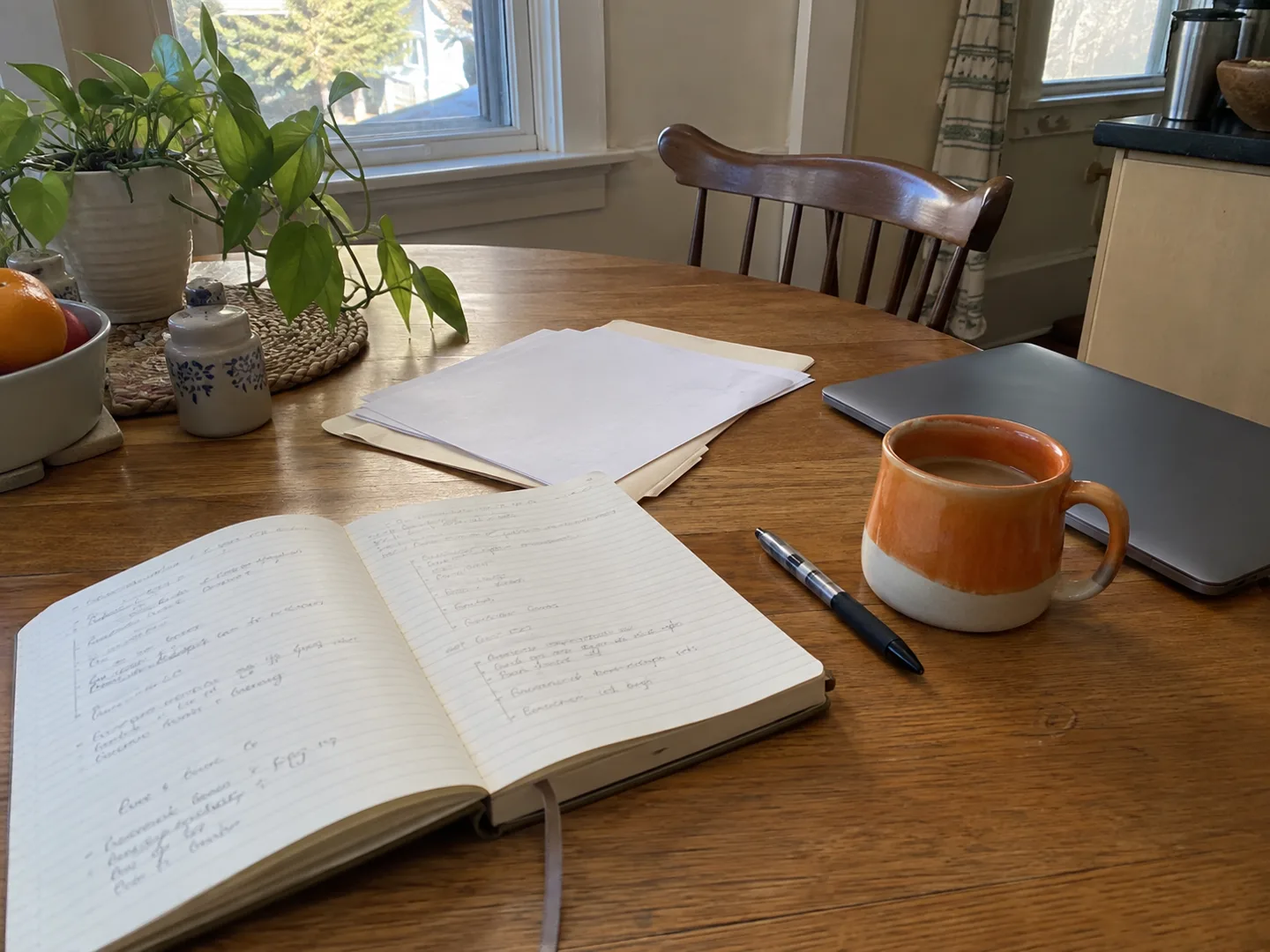 Notebook, paperwork, coffee and a laptop on a table in natural window light.